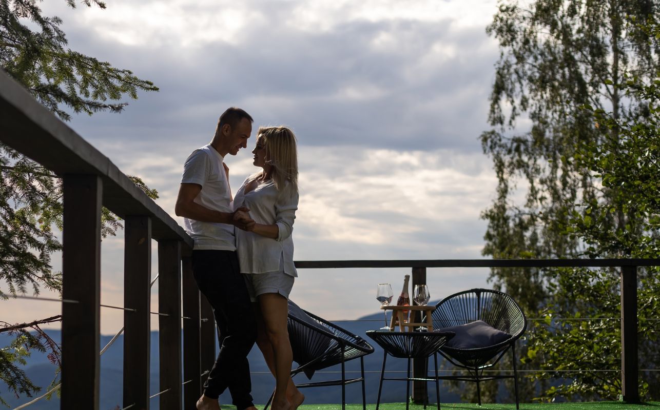 Romantic couple sharing a quiet moment on a scenic mountain-view deck with modern outdoor seating, wine glasses, and a bottle of rosé at sunset.
